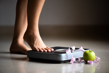 Female leg stepping on weigh scales with measuring tape and green apple. Healthy lifestyle, food...