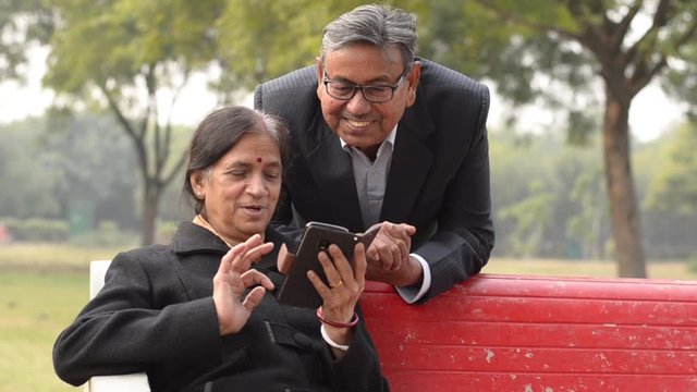 Portrait of a happy looking retired senior smart Indian man and woman couple smiling, talking and looking at mobile phone screen while sitting on a park bench in New Delhi, India. Concept love