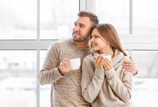 Happy couple with cups of hot tea resting near window at home