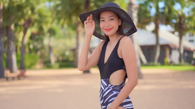 A pretty Asian woman in her bathing suit moves in the frame as she adjusts her floppy sun hat.