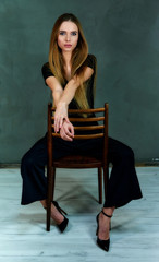 The model sits in different poses on a chair in front of the camera. The concept of glamor, style. Portrait of a pretty blonde girl with long hair and excellent make-up on a gray background.