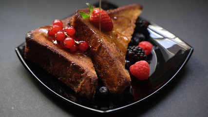 Plate of delicious French toast with many kinds of berries ( raspberries ,strawberries and blueberries ) and pouring honey or syrup on french toast , black table background , 