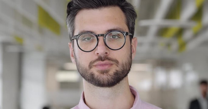 Close up of bearded caucasian man in 30s looking to camera and blinking. Portrait of handsome guy with serious face expression. Blurred modern office background. Concept of people.