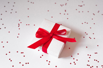 White gift box with red satin ribbon on white background with confetti. Background of Valentine's day, Christmas or New Year