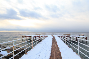 Winter beach. Blue clear sea water, snow and a wooden pier. Skyline and mountains.