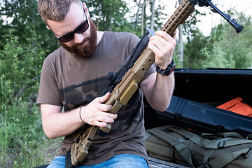 A man with glasses with a beard holds a sniper rifle
