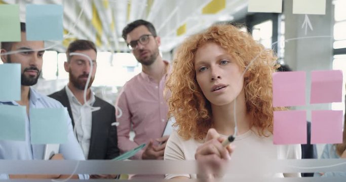 Close up of female office worker using glass board and proposing her problem solving to workmates. Diverse coworkers having corporate meeting and standing near glass wall with sticky notes.