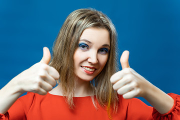 Beautiful young girl shows thumbs up on two hands. In a red dress on a blue background. The concept of a promise, like, support