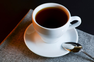 White cup of black tea served on a plate with a spoon on dark table background. Hot drink.