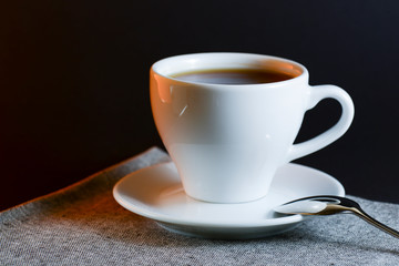 White cup of black tea served on a plate with a spoon on dark table background. Hot drink.