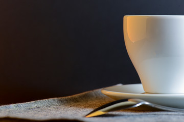 White cup of black tea served on a plate with a spoon on dark table background. Hot drink.