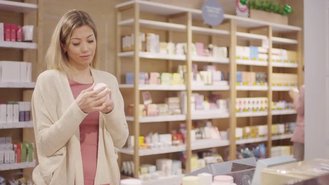 Tracking shot of cheerful young woman opening bottle of cream and putting it on her hand while choosing cosmetic product at store