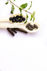 Black and brown ayurvedic pills in spoon, long pepper and tulsi leaves on white background. Aayurveda concept