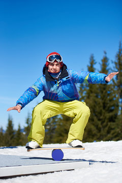 Smiling athletic male snowboarder training, balancing on snowboard on snowy hill on background of blue sky and spruce trees on sunny day. Winter extreme mountain sports, active lifestyle concept.