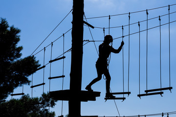 Silhouette of girl in helmet climbing on high rope course against blue sky