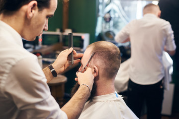 Barber servicing adult client in men's hairdressing salon. Coiffeur completing his working process with final movements of scissors. Focus on cutting hair with scissors and comb in barber's hands.