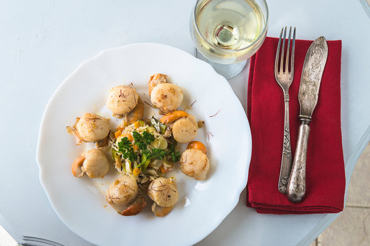 Gourmet concept. White plate with  st james shells or pecten maximus with saffron, vintage cutlery on the red napkin, glass of white wine on white table top view