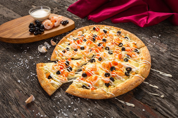 Pizza with very much cheese melting. A slightly pushed slice of fresh pizza with tomatoes, cheese, black olives, shrimp, sauce and garlic on old wooden table with dark red fabric
