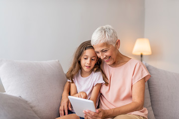 Smiling granddaughter and grandmother with tablet pc computer sitting on couch at home. Picture of cheerful young lady sitting at home with her grandmother using tablet computer.