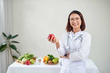 Smiling nutritionist in her office, she is holding a green apple and showing healthy vegetables and...