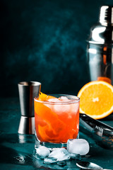 Orange Alcohol cocktail with red vermouth, bitter, soda, orange zest and ice, dark wooden bar counter background, bar tools, selective focus
