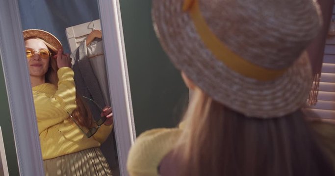 View over the shoulder on the Caucasian young pretty blonde girl fashion blogger in a hat looking in the mirror while trying on sunglasses. Indoors.