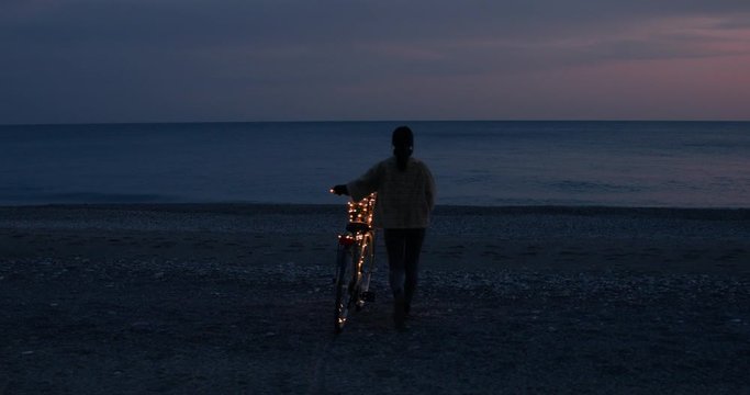 Ragazza va in giro in bicicletta di notte sulla spiaggia. Striscia illuminata di luci led.