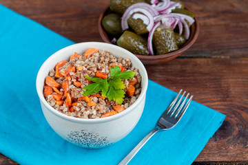 Buckwheat porridge in a bowl with stewed carrots. In the background are fermented pickled cucumbers and red chili peppers.