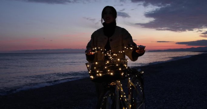 Sagoma di una ragazza in bicicletta che cammina in riva al mare di notte al tramonto. Striscia led.