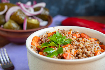 Buckwheat porridge in a bowl with stewed carrots. In the background are fermented pickled cucumbers and red chili peppers.