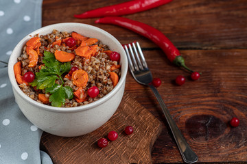 Buckwheat porridge in a bowl with stewed carrots. In the background are fermented pickled cucumbers and red chili peppers.