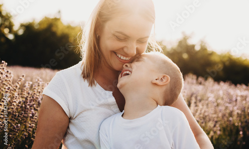 Portrait of a beautiful young mother laughing while holding on her legs her little kid which is laughing while closed eyes in a field of flowers.