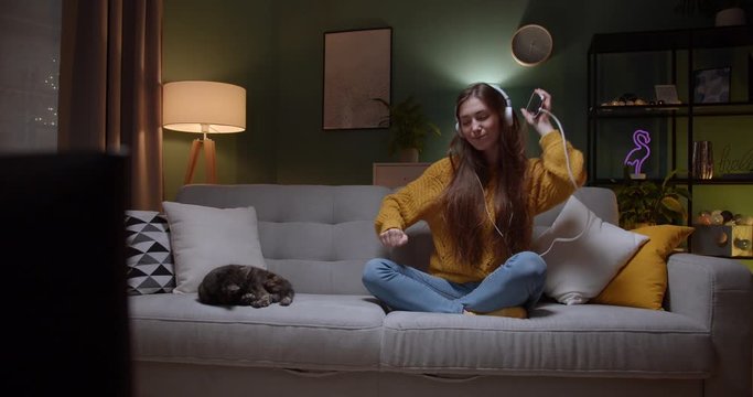Young Caucasian pretty girl in headphones sitting on the couch with a cat beside and dancing while listening to the music on the smartphone in the dark room at night. Indoor.