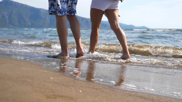 Legs of young couple stepping together along beach at ocean background. Pair walking near sea and splashing water washing their feet. Male and female legs going on sandy shore with waves. Close up
