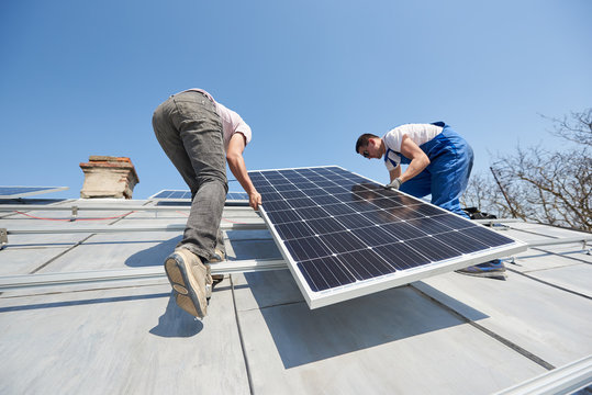 Male engineers installing residential solar photovoltaic panel system. Electricians lifting blue solar module on