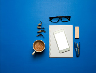 beautiful flat composition inspired by the color of the year 2020 (classic blue) white paper on top of white phone with fill screen, fir leaves, stapler, piece of wood black glasses & coffee 
