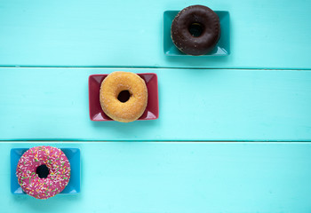 Three donuts with different fillings and icing on three rectangular saucers on a wooden blue background top view