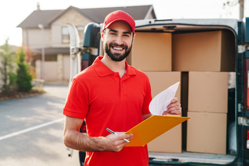 Image of cheerful delivery man holding clipboard and writing - obrazy, fototapety, plakaty