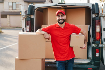 Image of joyful young delivery man standing with parcel boxes near car