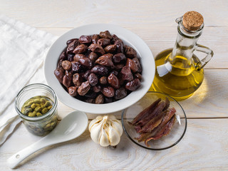 Ingredients for tapenade: dried or cured olives in a white bowl, anchovy fillet, capers, garlic and olive oil on a white rustic table. Mediterranean cuisine.