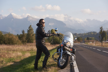 Bearded tall athletic motorcyclist in dark sunglasses, black leather clothing standing at modern powerful motorbike on background of green rural landscape, steep mountain peaks and bright sky.