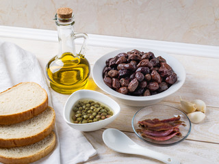 Ingredients for tapenade: cured or dried olives in a white bowl, anchovy fillet, capers, garlic, olive oil and bread on a white rustic table. Mediterranean cuisine.