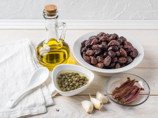 Ingredients for tapenade: cured or dried olives in a white bowl, anchovy fillet, capers, garlic and olive oil on a white rustic table. Mediterranean cuisine.