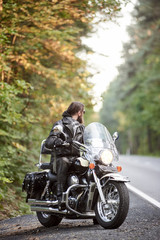 Obraz na płótnie Canvas Bearded biker in black leather clothing sitting on cruiser motorcycle on country roadside on background of empty straight asphalt road and green trees bokeh foliage.