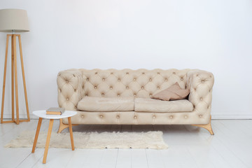 A beige sofa with a decorative pillow, a coffee table and a lamp in a spacious white living room. Spacious room interior with comfortable sofa against a white wall. Home decor. Scandinavian style 