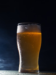 A glass of cold light beer lager on a dark background with smoke
