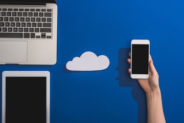cropped view of woman holding smartphone near empty white paper cloud and laptop with digital tablet on blue