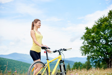 Naklejka premium Attractive happy girl cyclist standing on a grassy hill with yellow mountain bicycle, enjoying summer day in the mountains. Outdoor sport activity, lifestyle concept