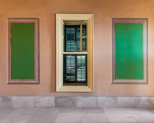 Narrow wooden window with closed green shutters mediating two beautiful elegant rectangular green frames on orange wall with white marble floor, in abandoned old building