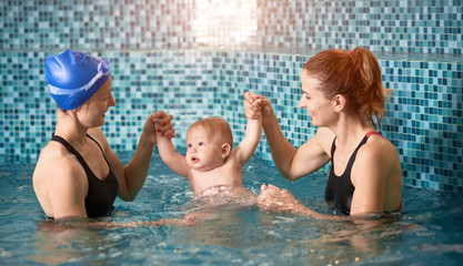Two women holding a baby boy hands up in swimming pool on blue background. Safety swim activities...